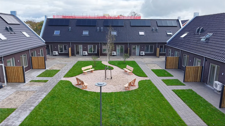Binnenplaats van moderne woningen met een centraal pad, groene gazons, houten banken en zonnepanelen op de daken.