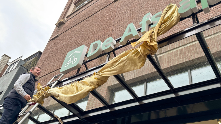 Een man op een ladder onthult samen met een goudkleurig doek een gevelbord met een fietslogo en de tekst 'De Afstap'.