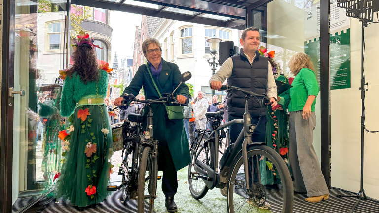 Twee mensen duwen hun fietsen door een glazen deur, met een publiek buiten zicht. Een vrouw in een groene jas en bril en een man in een zwarte bodywarmer zijn zichtbaar. Een vrouw met een groene jurk versierd met bloemen staat met haar rug naar de kijker. De omgeving geeft een stadscentrum weer met gebouwen en mensen op de achtergrond.