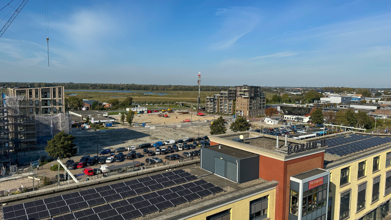 Bouwplaats met meerdere gebouwen in aanbouw, omgeven door parkeerterreinen en een weids landschap met water en bossen in de achtergrond onder een heldere blauwe lucht.