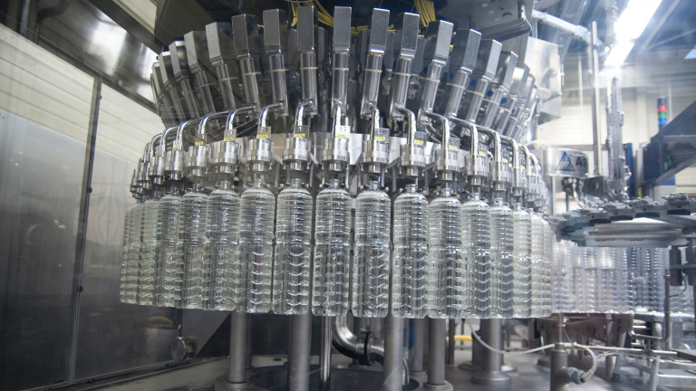 Rij met doorzichtige plastic flessen die door een industriële vulmachine worden gevuld in een fabriekshal.