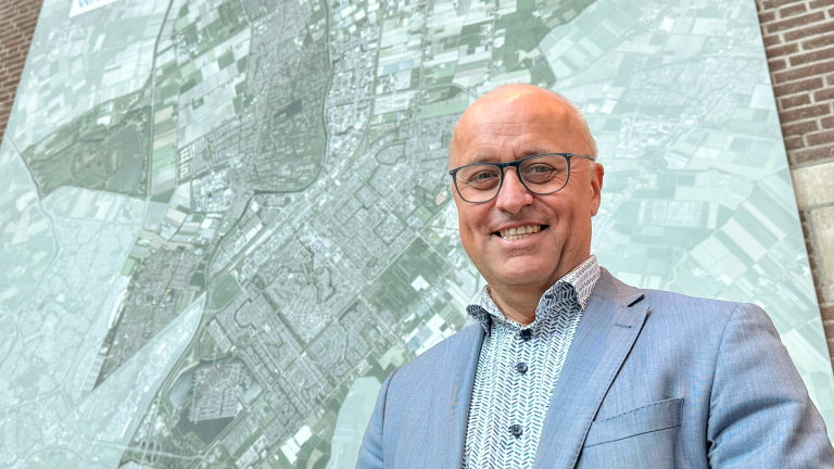 Man in a lichtblauw pak en bril glimlacht naar de camera, met een grote kaart van een stedelijk gebied op de achtergrond.