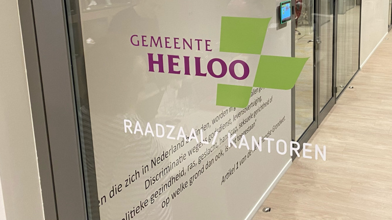 Glazen deur met daarop het logo en de tekst 'Gemeente Heiloo'. Onder het logo staan de woorden 'Raadzaal' en 'Kantoren'.