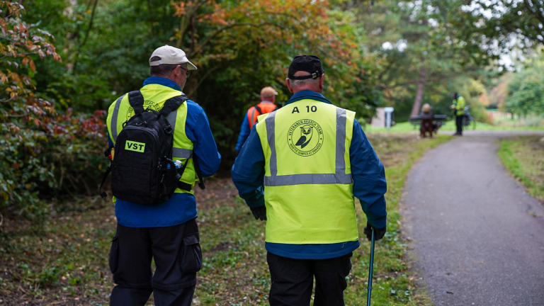 Twee mensen in gele veiligheidshesjes met rugzakken lopen op een pad in een groen park, met andere mensen in de verte.