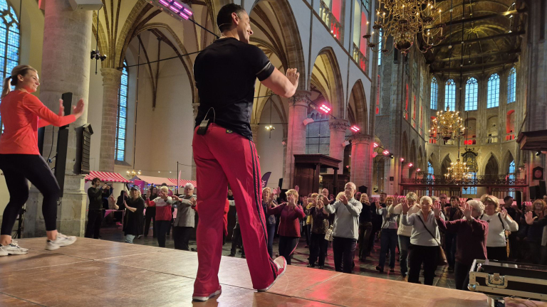 Een groep mensen voert een fitnessoefening uit onder begeleiding van twee instructeurs in een grote, historische kerk met hoge gewelven en kroonluchters.