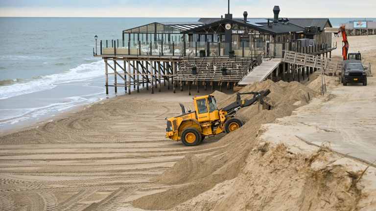 Een gele graafmachine is bezig met het verplaatsen van zand op een strand, naast een strandpaviljoen op palen aan de kust.