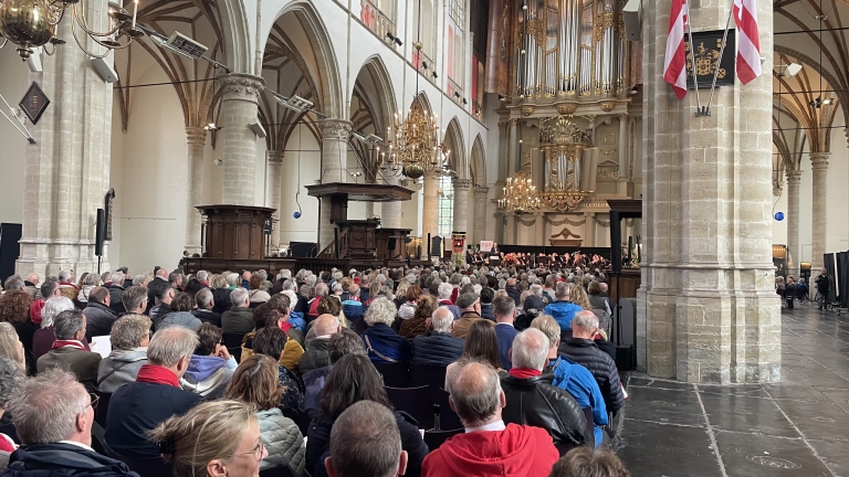 Een volle kerkzaal met veel mensen die op stoelen zitten en luisteren naar een optreden of toespraak. Het interieur bevat hoge gewelven, grote zuilen en een orgel. Er hangen kroonluchters en vlaggen in de ruimte.