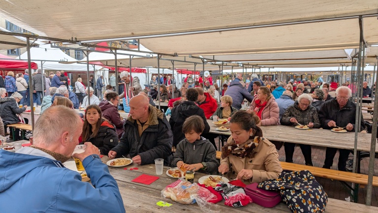Mensen zitten aan houten tafels onder tentdoeken op een drukbezocht buitenfestival, etend en pratend, met verschillende leeftijden zichtbaar.