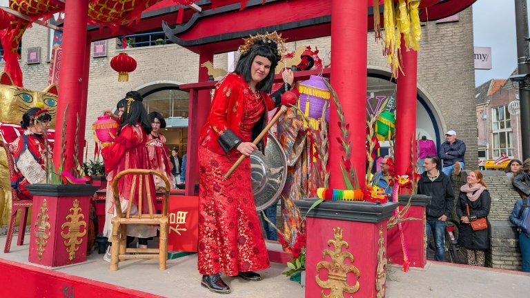 Mensen in traditionele kleding poseren op een sierlijke, rode wagen versierd met Chinese draken en een grote gouden kat tijdens een parade, terwijl omstanders toekijken.