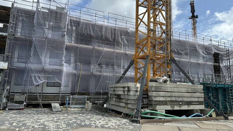 Bouwplaats met een hoog gebouw omgeven door steigers en netten, en een gele kraan op de voorgrond.