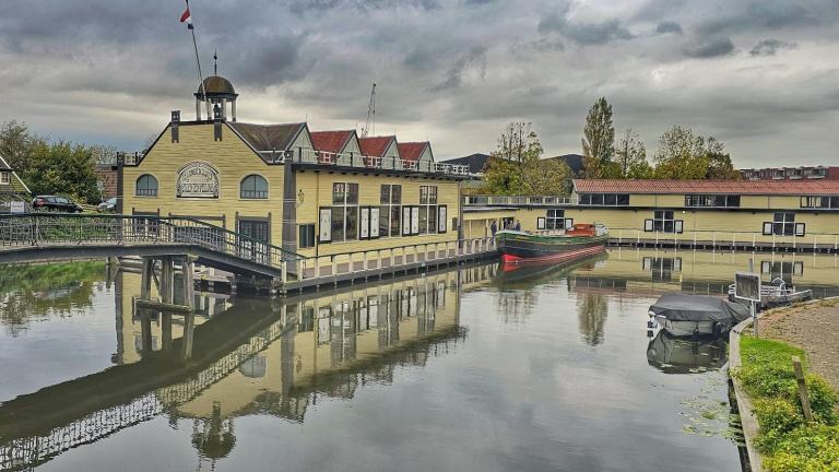 Geleen waterstraatscène met historische gebouwen aan het water, verbonden door een brug met geparkeerde boten en een vergrijsde lucht op de achtergrond.