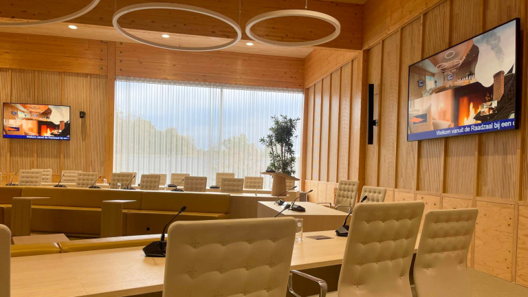 Vergaderzaal met houten panelen, grote ramen met gordijnen, tafels met microfoons, een podium met een plant, en schermen met afbeeldingen en tekst.