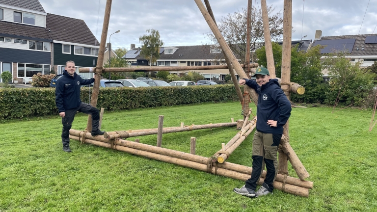Twee mensen in scoutingkleding poseren naast een grote houten constructie op een grasveld, met huizen en auto's op de achtergrond.