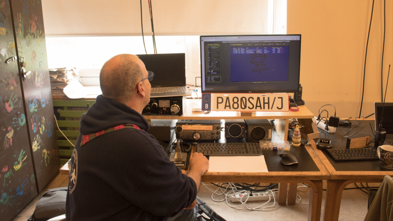 Een man in een donkere hoodie kijkt naar een computerscherm met radiocommunicatieapparatuur op een bureau. De ruimte heeft een kleurrijk beschilderde kast en een bord met het opschrift 'PA8ØSAH/J' staat prominent op het bureau.