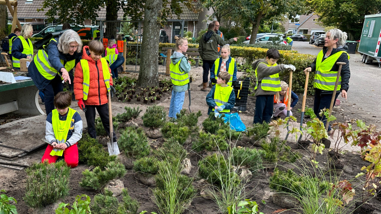 Mensen, voornamelijk kinderen, dragen gele veiligheidsvesten en planten groene struiken in een tuinomgeving.