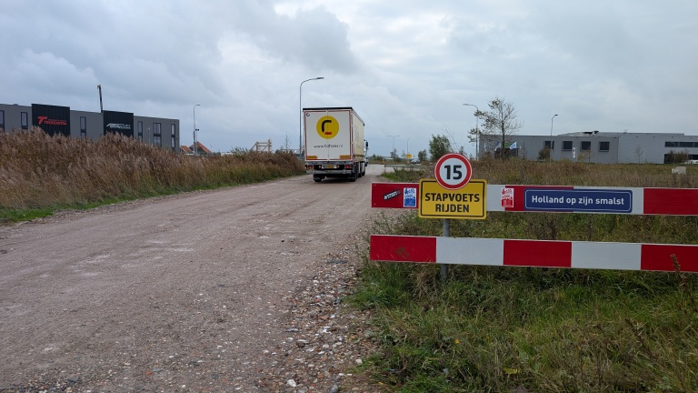 Een vrachtwagen rijdt over een onverharde weg naast hoge grassen, met links moderne gebouwen in de verte; op de voorgrond staat een bord met snelheidslimiet van 15 km/u en tekst "Stapvoets rijden" en "Holland op zijn smalst."