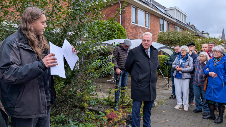 Een man met lang haar houdt enkele papieren vast terwijl hij buiten spreekt tot een groepje mensen die aandachtig luisteren, naast een gebouw.
