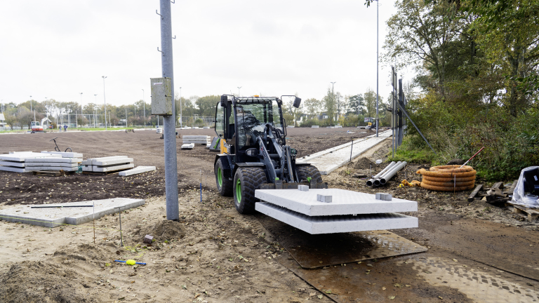 Een kleine wiellader vervoert betonnen platen op een bouwplaats met zandgrond en stapels platen op de achtergrond, omgeven door bomen en gereedschap zoals buizen en kabelrollen.