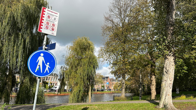 Park met borden, waaronder een blauwe wandelbord en een bord met gedragsregels, omgeven door bomen naast een rivier.