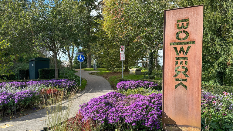 Parkpad met bloemen en groene bomen, een roestbruine sculptuur met inscriptie "BOWEREK" aan de zijkant, en een wandelpad bord met tekst "Munnikenblik."