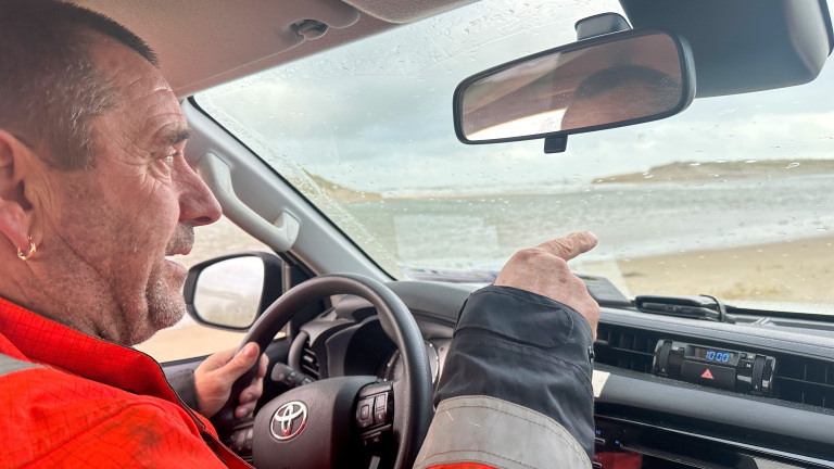Een man in een rode jas bestuurt een auto en wijst naar iets buiten het raam, terwijl het regent.