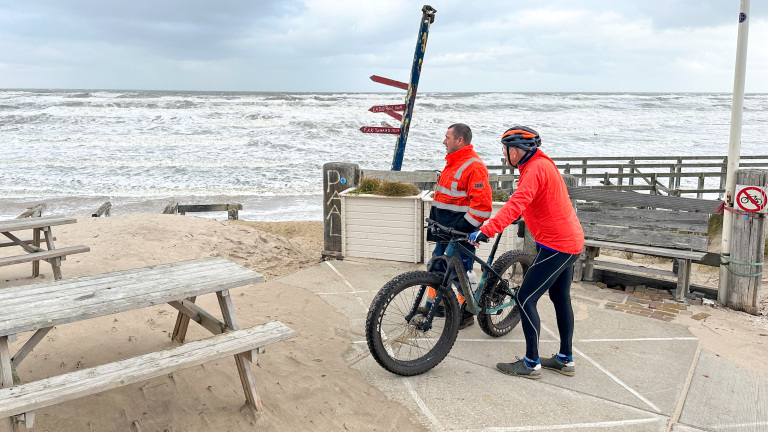 Twee mensen in felgekleurde kleding, waarvan één met fiets, kijken richting een woelige zee op een winderig strand. Een houten bank en verkeersbord zijn zichtbaar op de voorgrond.