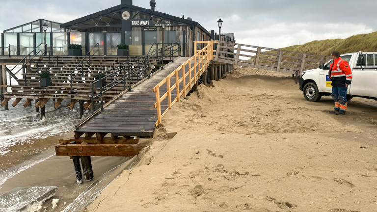 Een strandpaviljoen op stelten met een houten trap die naar het zand leidt, terwijl de golven eronder door spoelen. Een witte pick-uptruck staat geparkeerd in het zand, en een man in een oranje veiligheidsjas kijkt naar het gebouw.