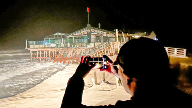 Iemand maakt 's nachts een foto van een strandpaviljoen nabij de woeste zee, met de silhouet van een persoon die een smartphone vasthoudt in de voorgrond.