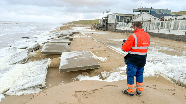 Een persoon in een oranje veiligheidsjas staat op een strand, waar grote betonnen platen door de branding zijn verschoven. Op de achtergrond zijn gebouwen zichtbaar die langs de kustlijn staan.