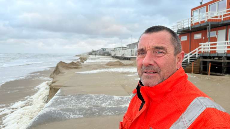 Een man in een feloranje jas staat op een strand, met een achtergrond van de zee en houten strandhuizen.