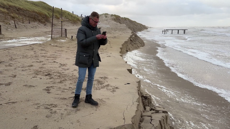 Man in winter clothing stands on eroded beach near the sea, taking a photo with a smartphone.