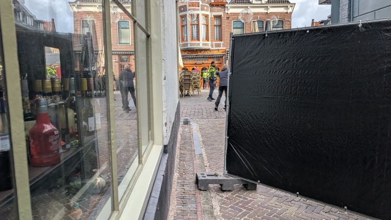 Straatscène met een zwart scherm en een etalage met flessen, terwijl op de achtergrond mensen en politie op een geplaveide straat bezig zijn voor een café met bruine gevels.