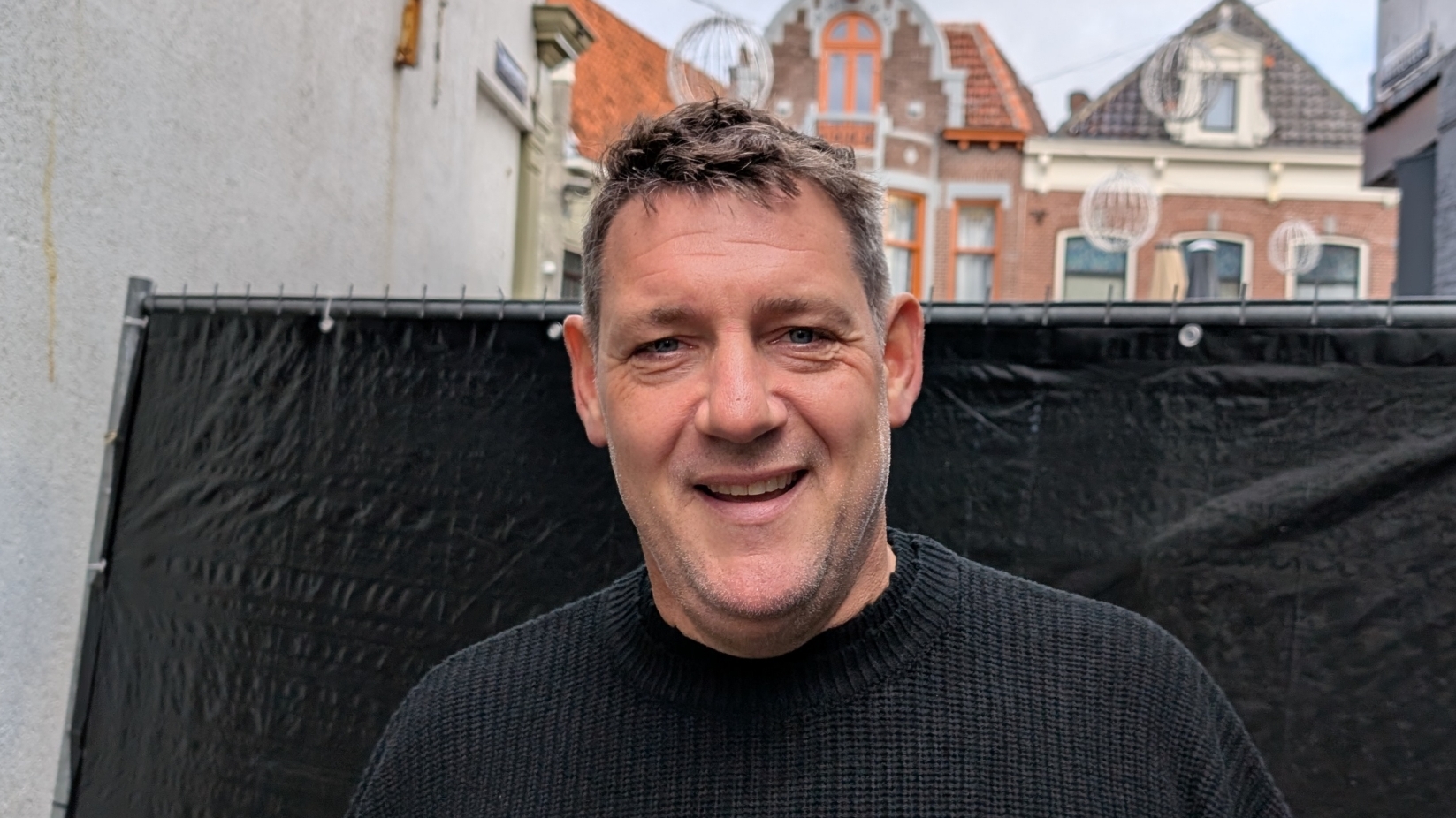 Man met een zwarte trui glimlacht naar de camera, met een achtergrond van oude gebouwen en een bouwhek.