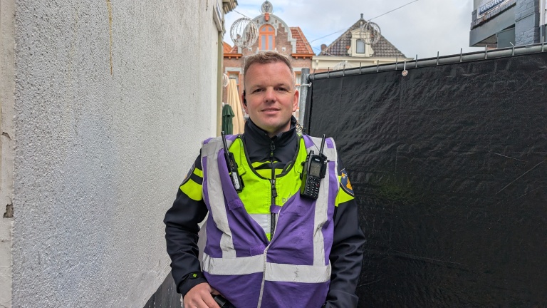 Politieagent in uniform met paarse veiligheidshes staat in een smalle straat, met historische Nederlandse gebouwen op de achtergrond.