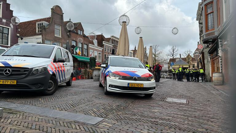 Een straat met twee politievoertuigen, een Mercedes-bus en een Volkswagen-auto, geparkeerd op een geplaveide weg naast enkele omstanders en politieagenten in uniform. De straat is omringd door historische gebouwen en versierselen hangen boven de weg.