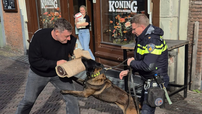 Een politieagent met een politiehond voert een trainingssessie uit met een man die een beschermende mouw draagt, met op de achtergrond een vrouw die toekijkt bij een etablissement met de naam 'Koning'.