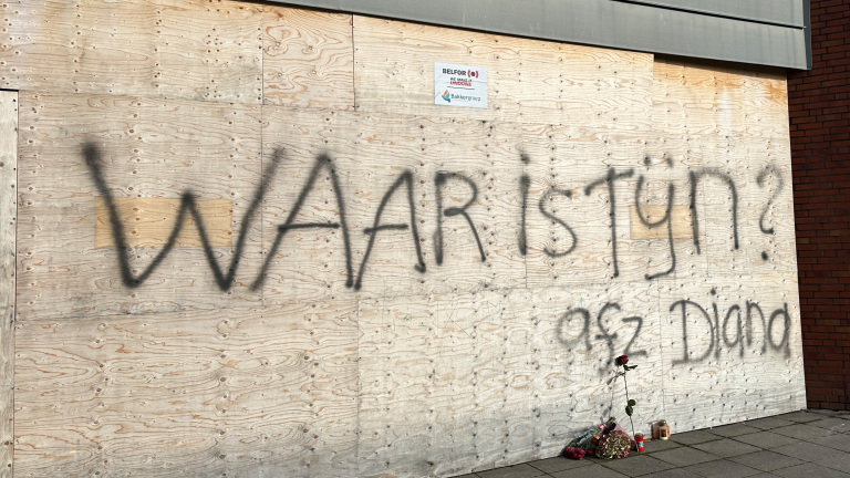 Op een houten muur is met graffiti de tekst "WAAR is Tijn? 9st² Diana" geschreven; er liggen bloemen en kaarsen op de grond.