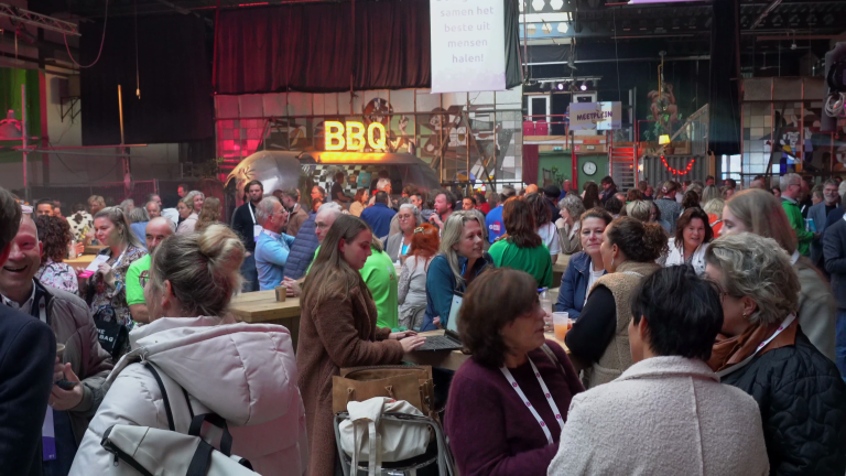 Drukke bijeenkomst met mensen die in groepjes staan te praten en lachen in een grote, sfeervolle ruimte, met een verlichte BBQ-signage op de achtergrond en een bar met mensen die drankjes bestellen.