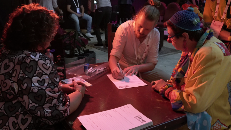 Een groep mensen zit aan een tafel waarvan één persoon een document ondertekent, terwijl anderen toekijken in een zaal met een informele sfeer.