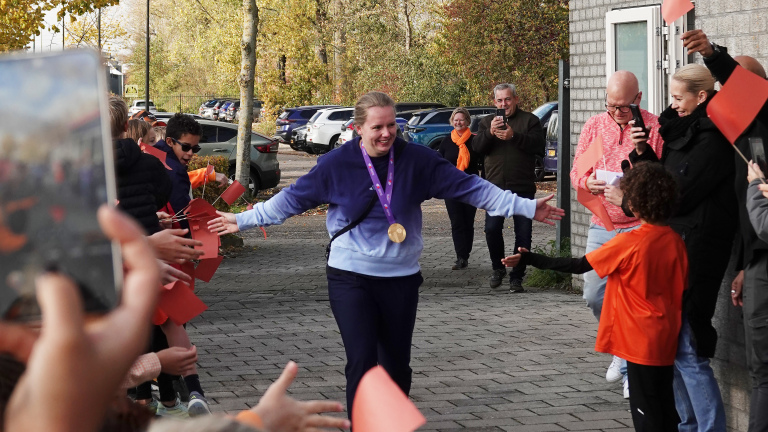 Vrouw met medaille loopt lachend door een rij mensen buiten, waarbij ze handen schudt terwijl ze wordt toegejuicht.