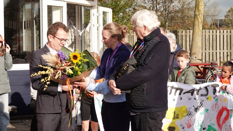 Een persoon in een pak ontvangt een bloemenboeket van een vrouw tijdens een buitenbijeenkomst, terwijl kinderen en andere volwassenen toekijken. Achter hen hangt een versierde banner met kindertekeningen en een olympisch thema.