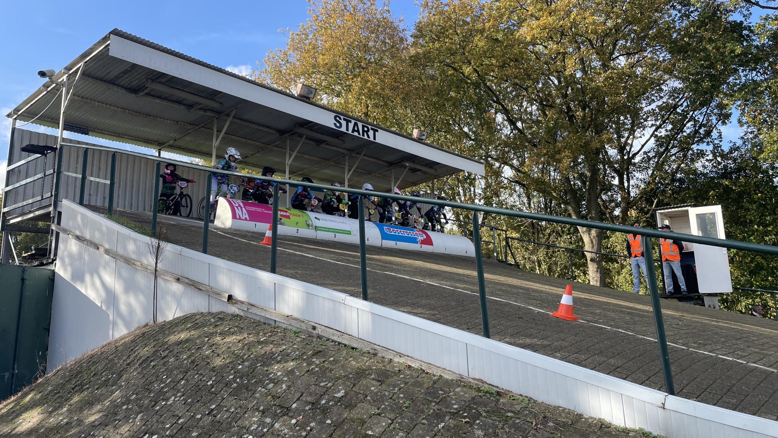 Startplatform van een BMX-race met rijders klaar op de baan en official in oranje hesje bij het startpunt, omgeven door bomen en blauwe lucht.