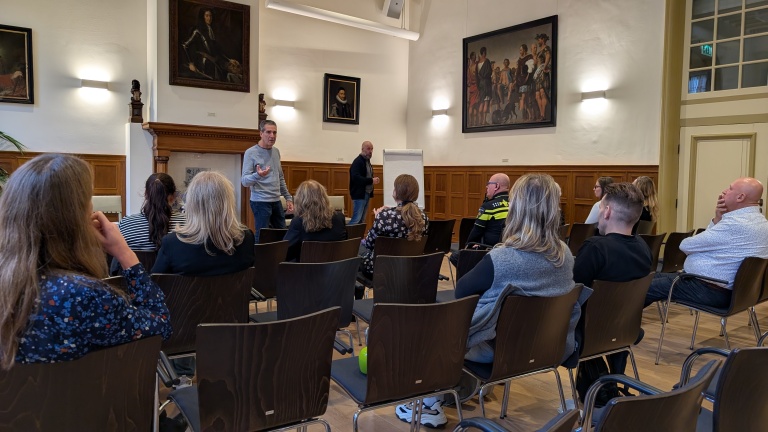 Mensen zitten op stoelen in een zaal met schilderijen aan de muur, terwijl een spreker voor de groep staat.