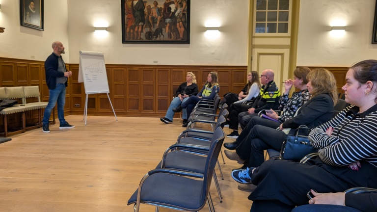 Een man presenteert aan een groep mensen die op stoelen zitten in een zaal met houten wanden, schilderijen en een flip-over.