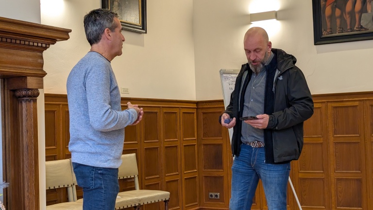 Twee mannen staan in een kamer met houten lambrisering aan de muur; de ene man houdt een smartphone vast terwijl de andere aandachtig luistert.
