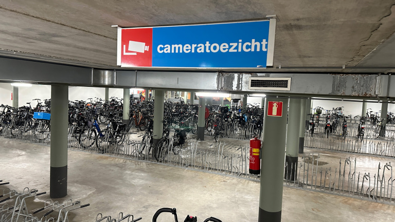 Ondergrondse fietsenstalling met veel geparkeerde fietsen en een bord met de tekst 'cameratoezicht' erboven, naast een blusapparaat aan een pilaar.