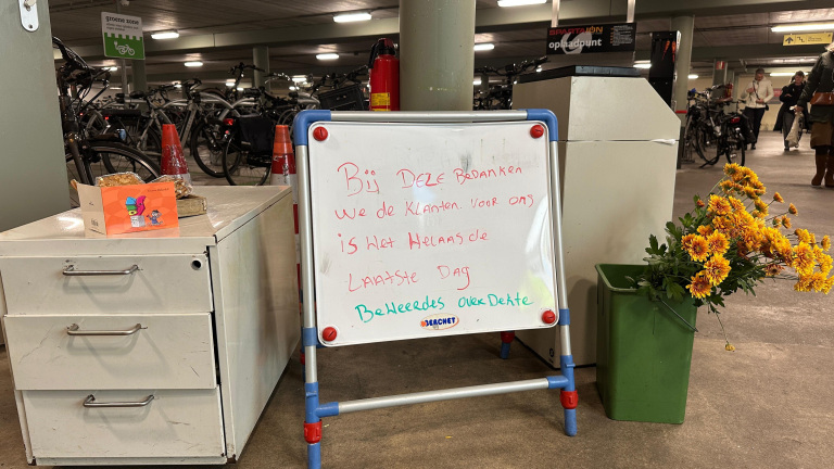 Een wit bord met rode en groene tekst staat naast een kastje en een groene emmer met bloemen in een fietsenstalling. Het bord vermeldt dat het hun laatste dag is en bedankt klanten.