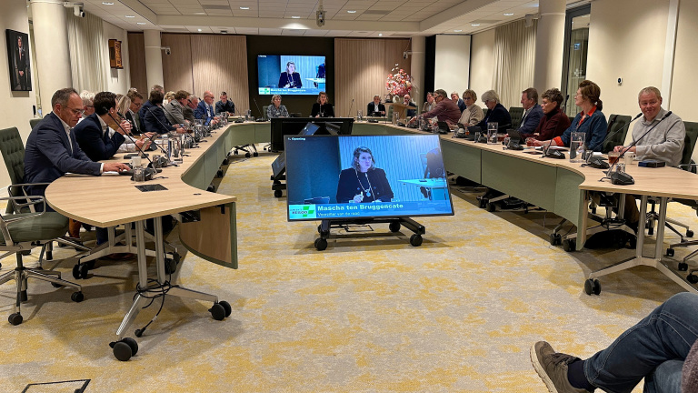Vergaderzaal met een groep mensen zittend aan een lange, gebogen tafel, kijkend naar een scherm waarop een vrouw te zien is met de tekst 'Mascha ten Bruggencate' en 'Voorzitter van de raad'.