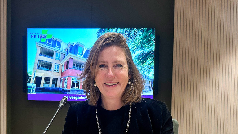 Vrouw glimlachend voor een microfoon met een scherm op de achtergrond waarop een gebouw en de tekst 'Gemeente Heiloo' te zien zijn.