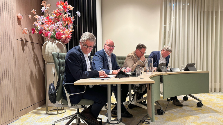 Vier mannen in formele kleding zitten aan een tafel met elektronische apparaten en papierwerk, met een grote bloemenvaas op de achtergrond in een vergaderzaal.